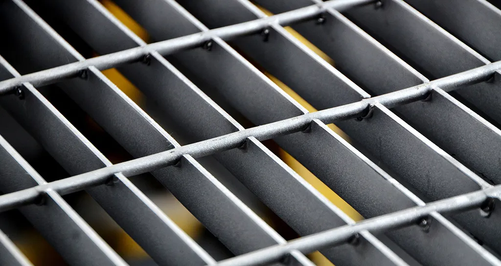 Steel bar grating — Macro detail of weld points on galvanized bearing bars; shallow depth of field; blurred machinery background