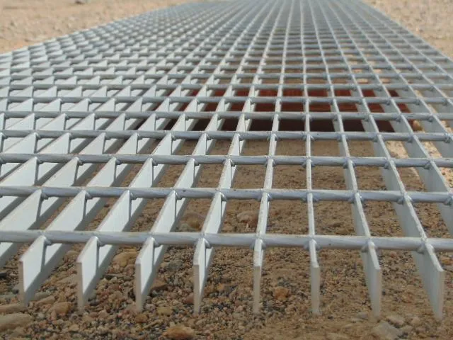 Welded steel grating close-up at ground-level angle showing bearing bars with round cross rods and visible weld joints; sandy/gravel ground setting