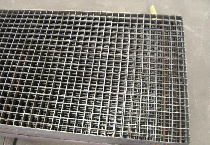 Stack of welded steel grating panels with twisted square cross bars on concrete factory floor; some surface rust