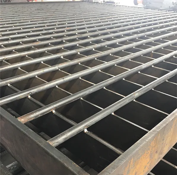 Bare welded steel grating panels stacked on factory floor; low oblique angle across bearing bars; machinery in background