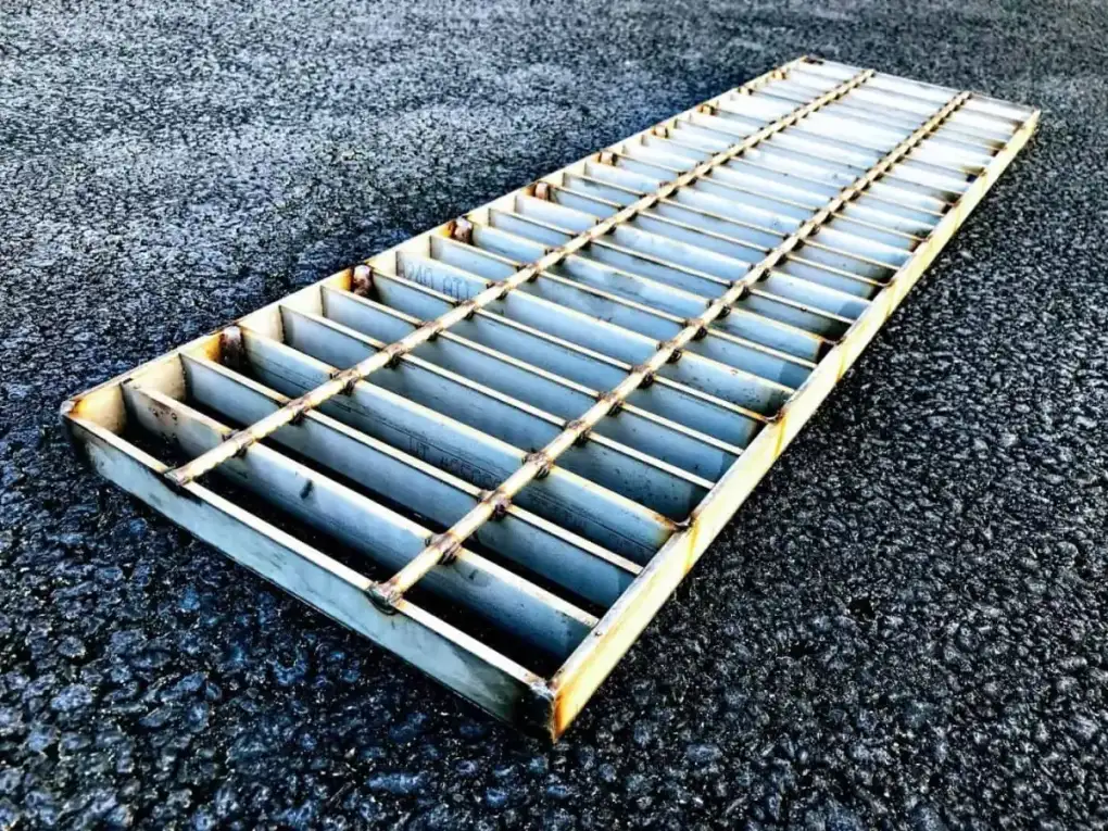 Welded steel grating panel resting on asphalt at low oblique angle; outdoor hard light